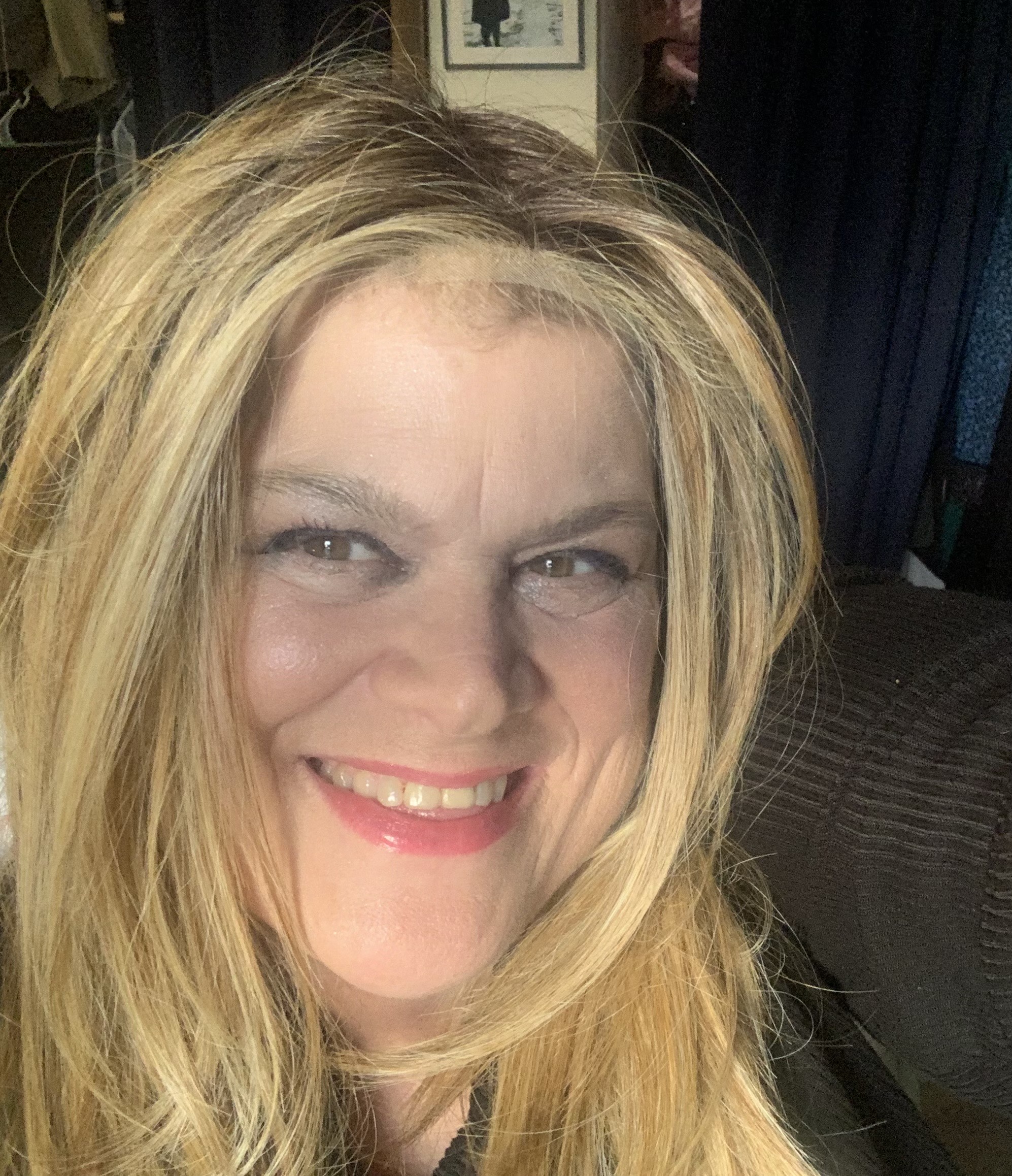 A woman with long, blonde hair smiles warmly at the camera, showcasing her bright lipstick and expressive eyes in a cozy indoor setting.