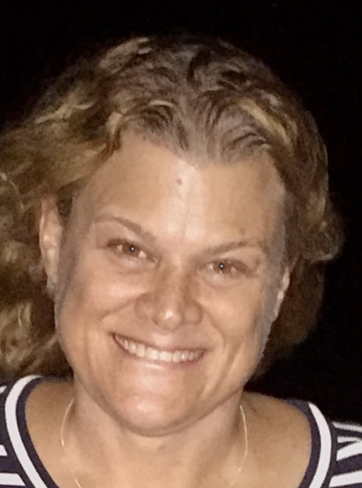 Smiling woman with curly hair, wearing a striped shirt, illuminated against a dark background. The image conveys a warm, friendly atmosphere.