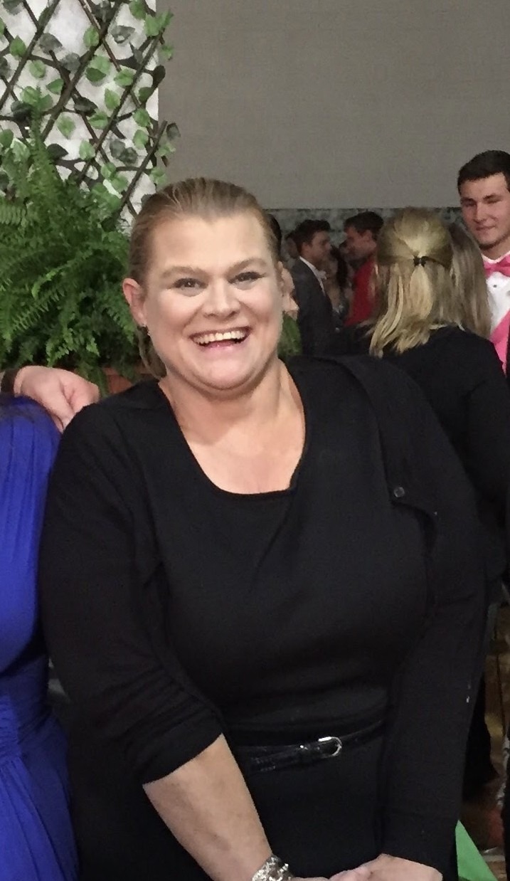 Smiling woman in a black dress stands amidst a festive gathering, with greenery in the background, embodying a joyful celebration.
