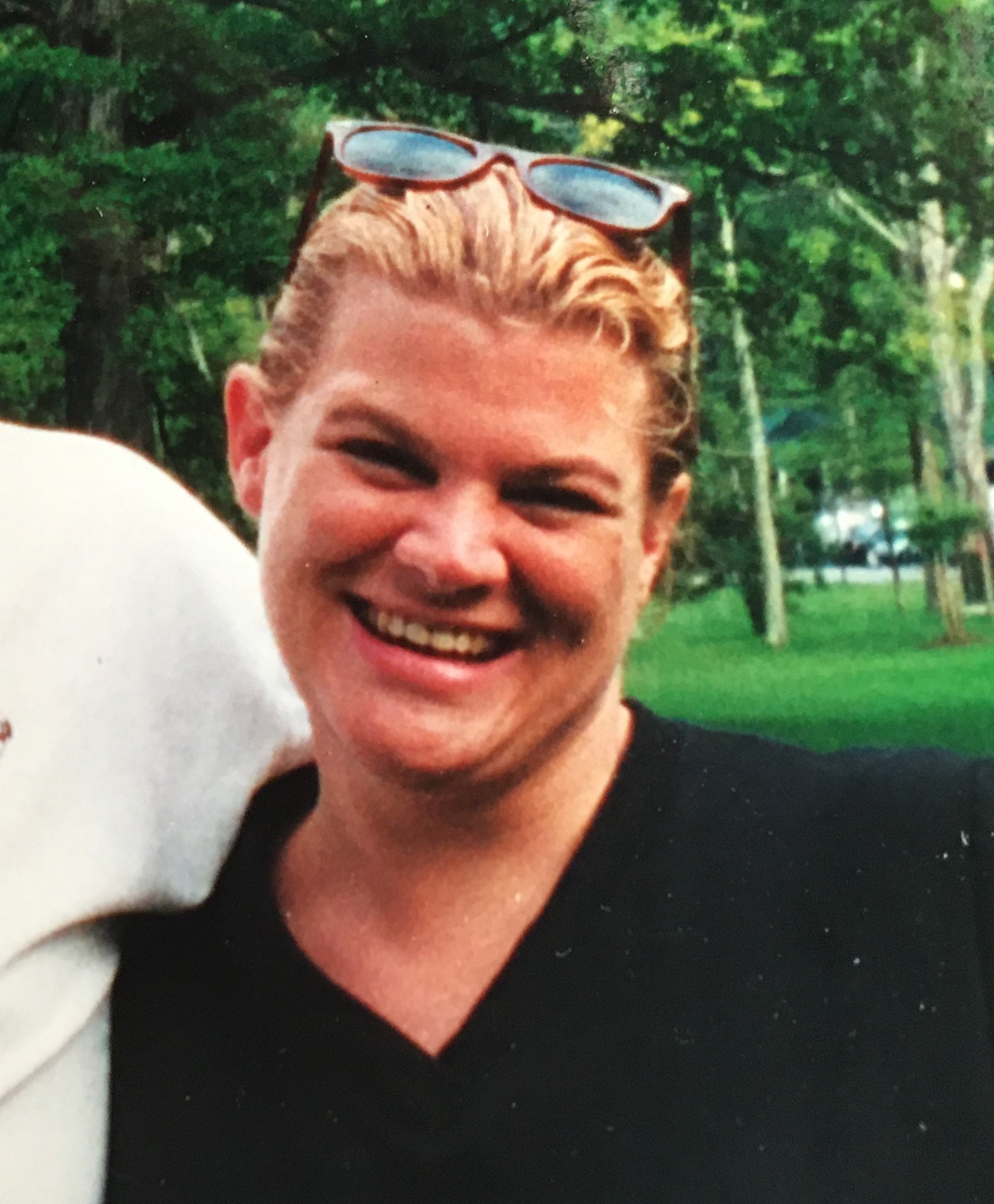 Smiling woman with blonde hair wearing a black shirt and sunglasses atop her head, standing outdoors amidst lush green trees.