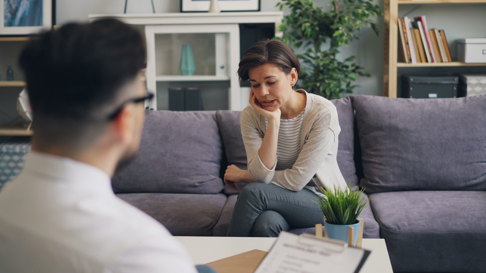 A woman in casual attire sits on a couch, looking contemplative during a counseling session, while a man in a white shirt listens attentively.